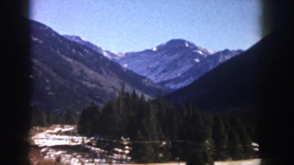 8mm film frame, c. 1961, Aspen area, Colorado: forested mountain slopes with snow patches in the valley and snow-capped peaks above under a clear blue sky.