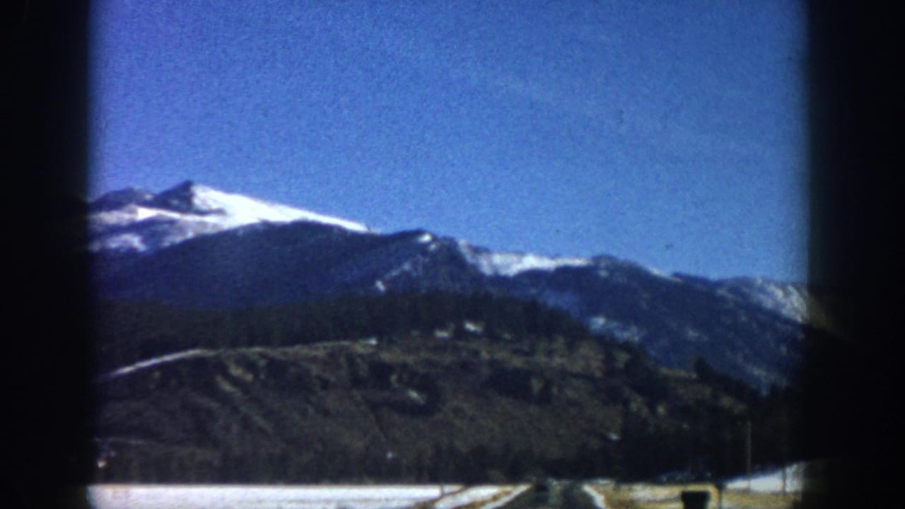 8mm home-movie frame, likely Aspen-area Colorado, 1961 — snow-capped peaks above dark conifer slopes with flat valley floor and road visible at bottom.