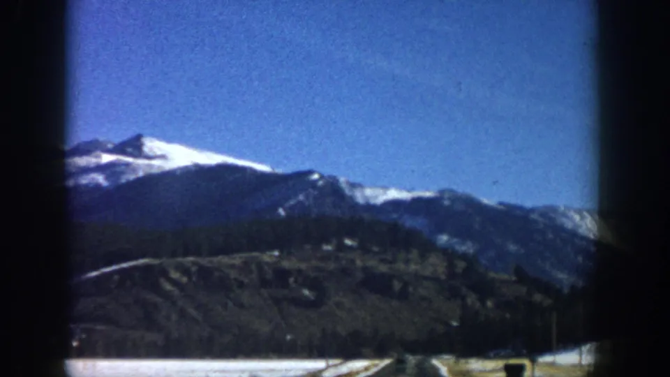 8mm home-movie frame, likely Aspen-area Colorado, 1961 — snow-capped peaks above dark conifer slopes with flat valley floor and road visible at bottom.