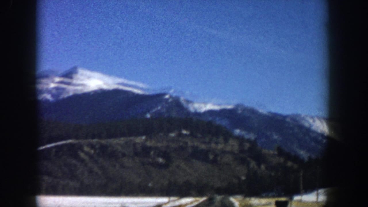 Prominent snow-capped peak with dark forested slopes and valley floor, film gate borders visible on sides.