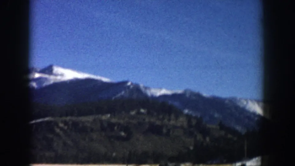 Final 8mm road-trip frame, Colorado mountain landscape, c.1961 — snow-capped peaks above conifer slopes with open valley floor.