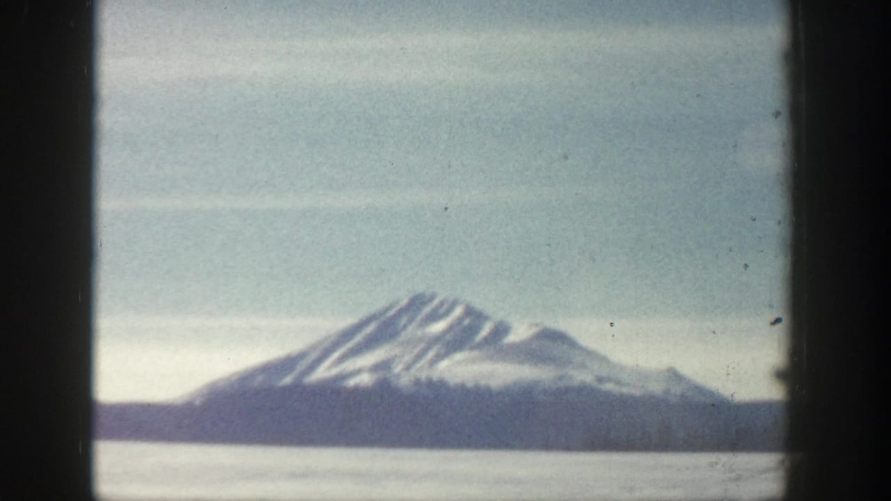 8mm home-movie frame from what may be Aspen, Colorado circa 1961, showing a snow-capped twin-summit peak above a flat valley under a pale blue sky.