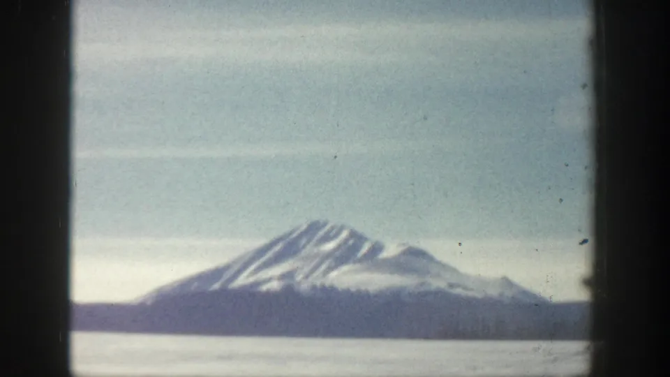 8mm home-movie frame from what may be Aspen, Colorado circa 1961, showing a snow-capped twin-summit peak above a flat valley under a pale blue sky.