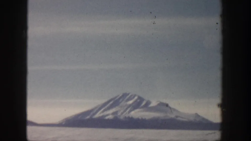 8mm home-movie frame, possibly Aspen, Colorado area circa 1961, showing a receding mountain peak with a broad snowy plain and overcast sky.
