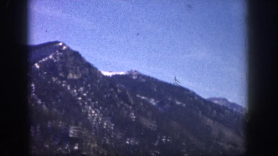 8mm home-movie frame, likely Colorado mountains circa 1961; a rocky ridgeline with snow fills the frame under a blue-purple color-shifted sky from film aging.