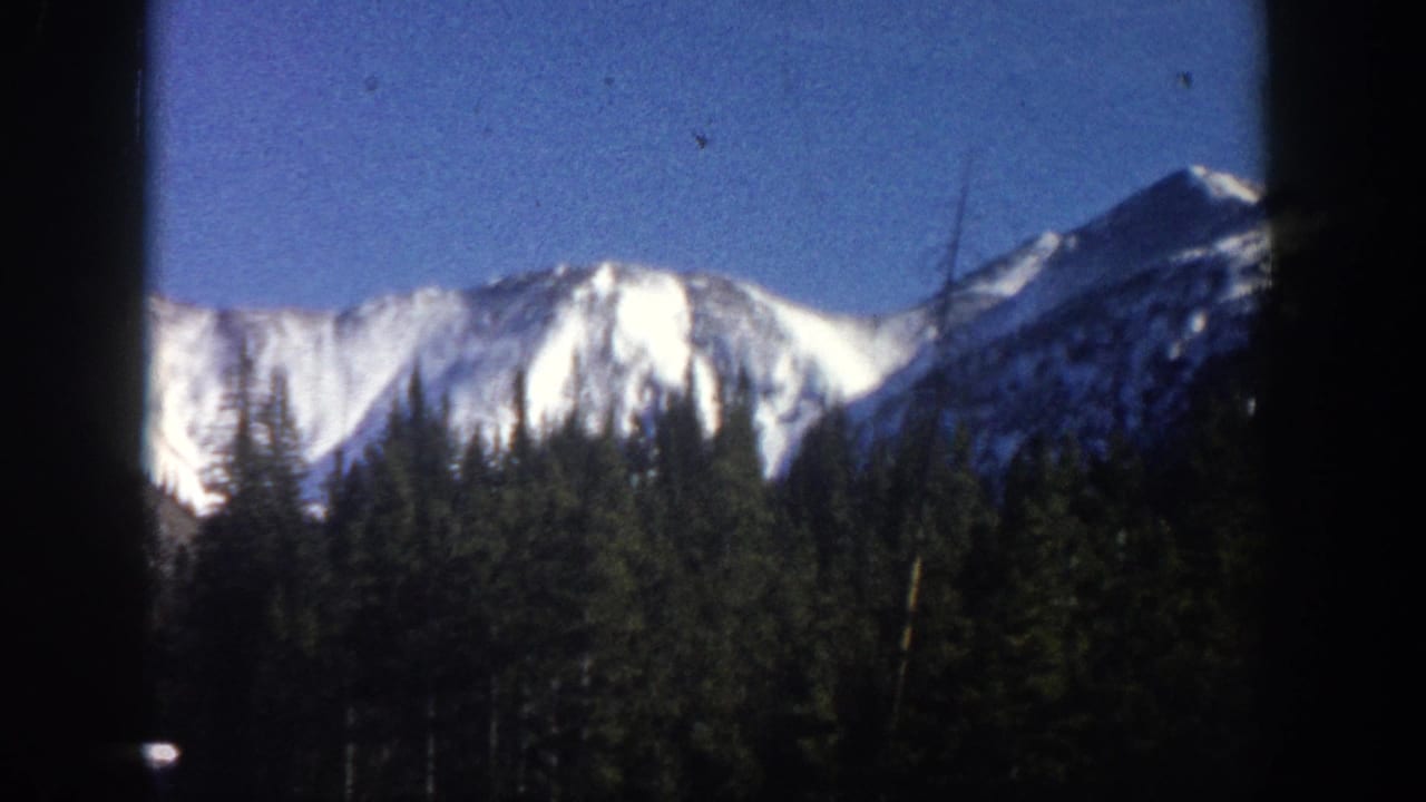 8mm home movie frame, Aspen Colorado area 1961 — snow-covered mountain peaks above dark evergreen forest under clear blue sky, with film grain and vignette borders.