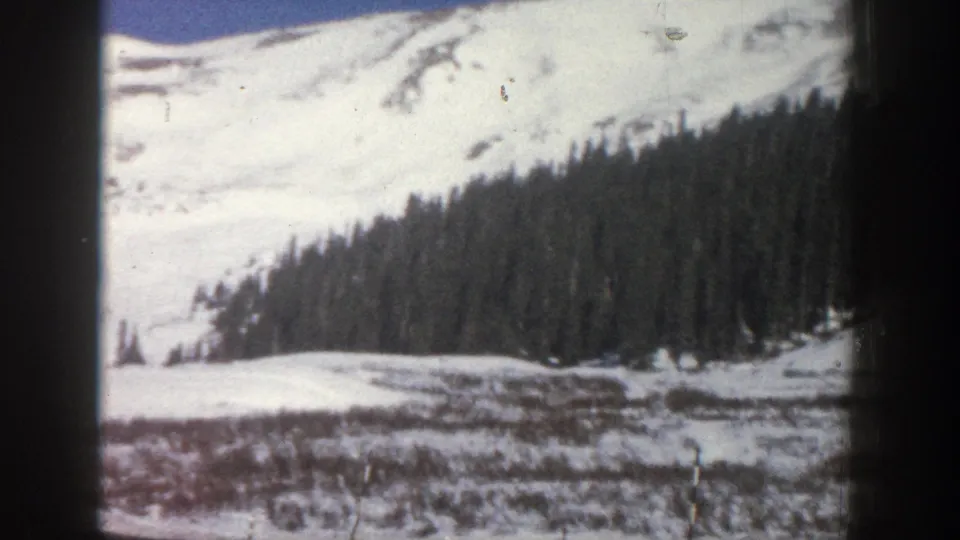 8mm home movie, Aspen Colorado area 1961 — wide snow slope with low scrub vegetation, evergreen forest at right, and partial mountain summit, shot from a moving vehicle.