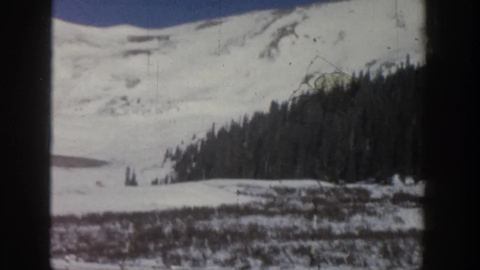 8mm home movie, Aspen Colorado area 1961 — snow slope with reddish scrub brush and sparse conifers in mid-distance; film scratches and grain are clearly visible.