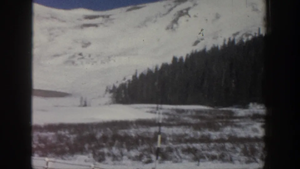8mm home movie, Aspen Colorado area 1961 — snow slope with scrub vegetation and scattered conifers; a thin vertical post or marker is visible at the right frame edge.