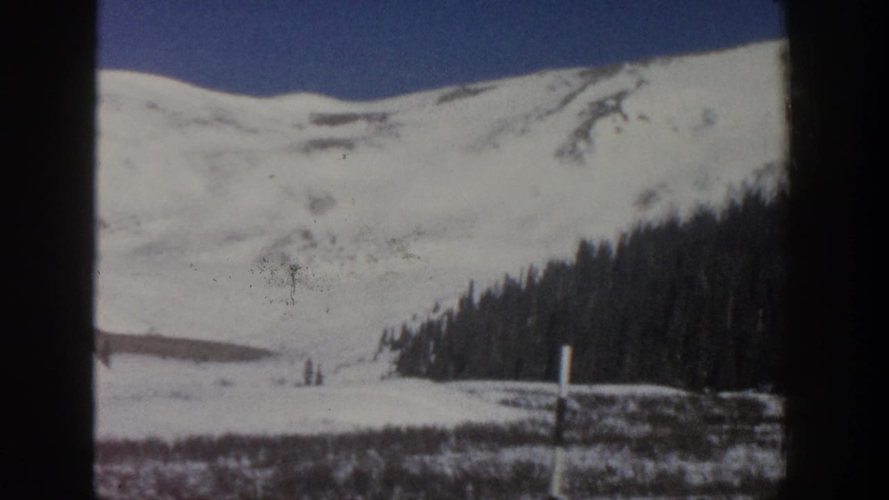 1961 Aspen, Colorado 8mm frame showing a snowy mountain ridge behind a dark evergreen forest and open blue sky.