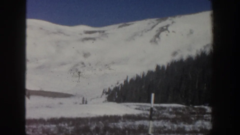 1961 Aspen, Colorado 8mm frame showing a snowy mountain ridge behind a dark evergreen forest and open blue sky.