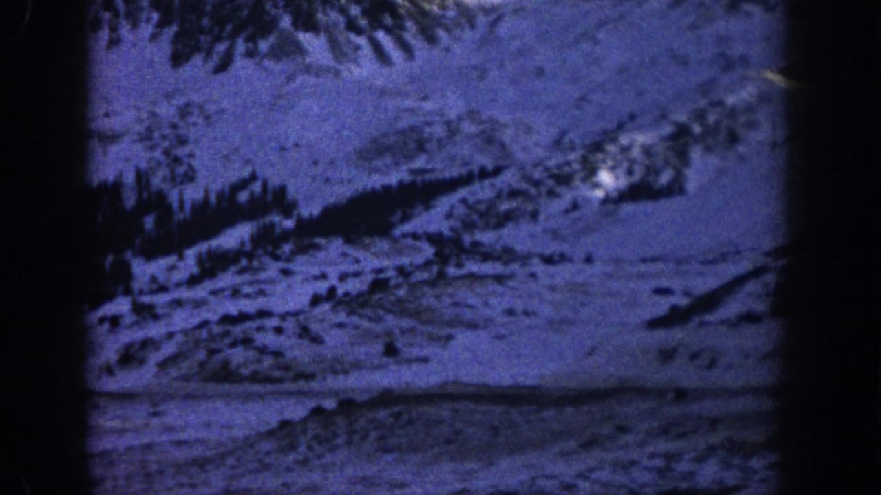 Blue-tinted 8mm view of snowy Colorado mountain terrain with evergreen trees and a road cutting through the slope.