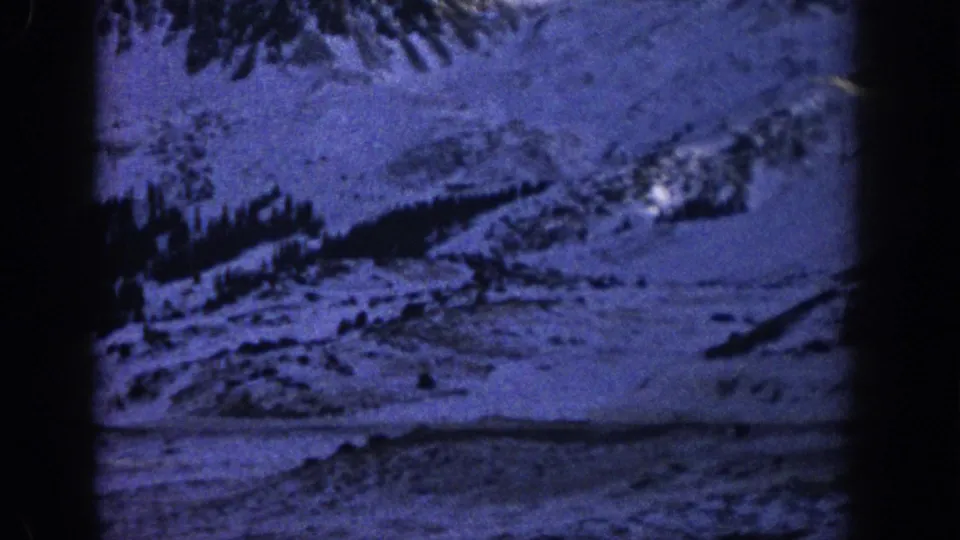 Blue-tinted 8mm view of snowy Colorado mountain terrain with evergreen trees and a road cutting through the slope.