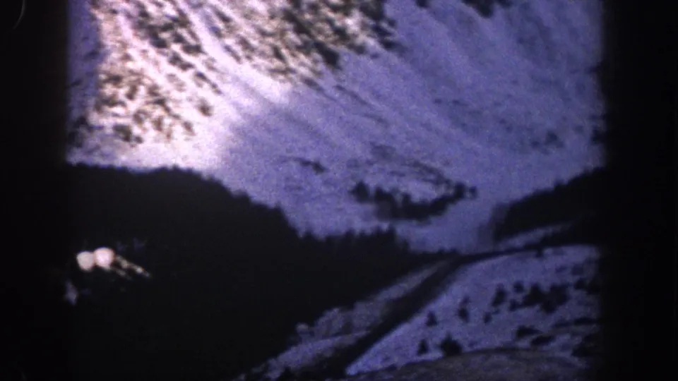 Low-light 8mm Colorado mountain view with a winding road, snowy hillside, trees, and a small distant light.