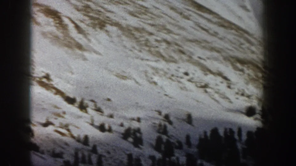1961 Aspen-area 8mm frame showing a snow-covered mountain slope with dark evergreen trees and patches of exposed earth.
