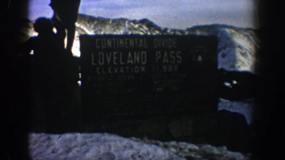 Visitors stand near the Loveland Pass Continental Divide sign in snowy Colorado mountains, seen in dark silhouette on 8mm film.