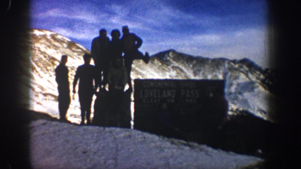 A small family group poses beside the Loveland Pass sign with snowy ridges and blue sky behind them in archival 8mm footage.