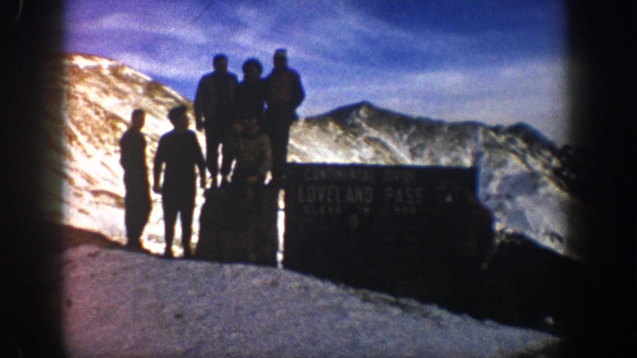 A 1961 home-movie frame shows several men posed at Loveland Pass, with the sign, snow, and high mountain backdrop visible.