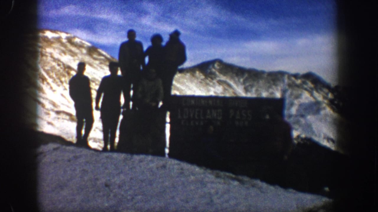 1961 Colorado 8mm frame of hikers silhouetted beside a Loveland Pass sign against snow-covered mountains and blue sky.