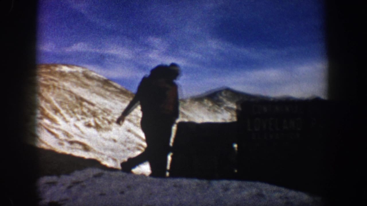 Silhouetted person in winter attire at an elevation marker, likely Loveland Pass, Colorado, circa 1961, rocky snow-covered peaks behind.