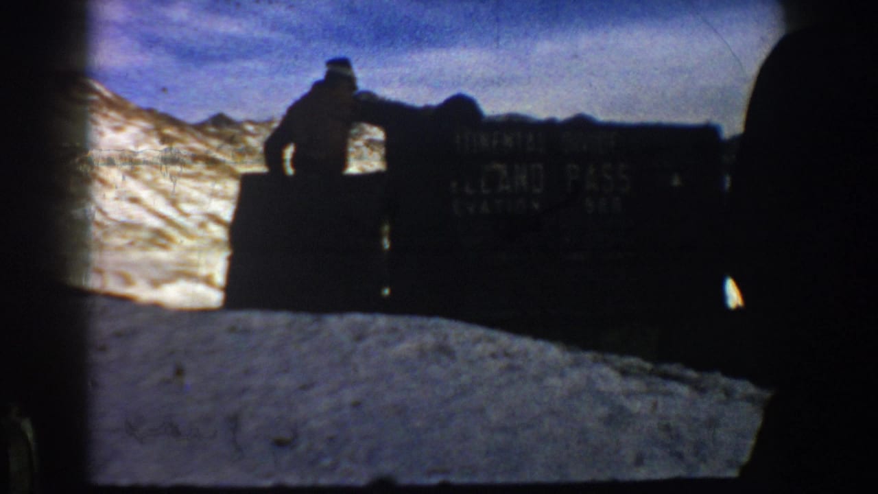 Two silhouetted figures near a large stone pass elevation marker, bright snow-covered mountains behind, blue sky.