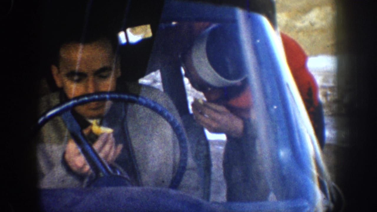 Young man eating while seated at the wheel of a 1960s-era car, blue steering wheel prominent, second passenger in red top visible at center.
