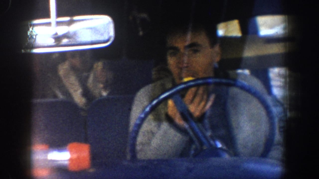 Young man at the wheel of a 1960s car eating during a road trip; rearview mirror and second figure visible in background.