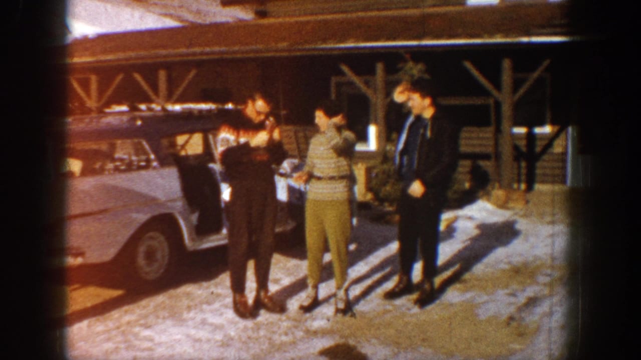 Three adults in early-1960s winter casual wear gathered beside a pale American sedan, timber-frame lodge structure visible behind them, Aspen, Colorado, 1961.