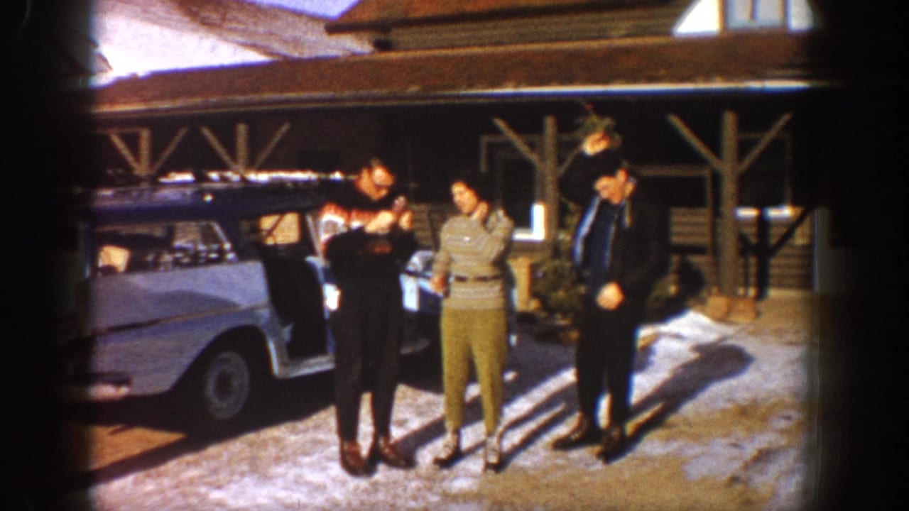 Three adults near a pale 1960s car, one in a patterned sweater, timber-frame structure and snow visible in background.