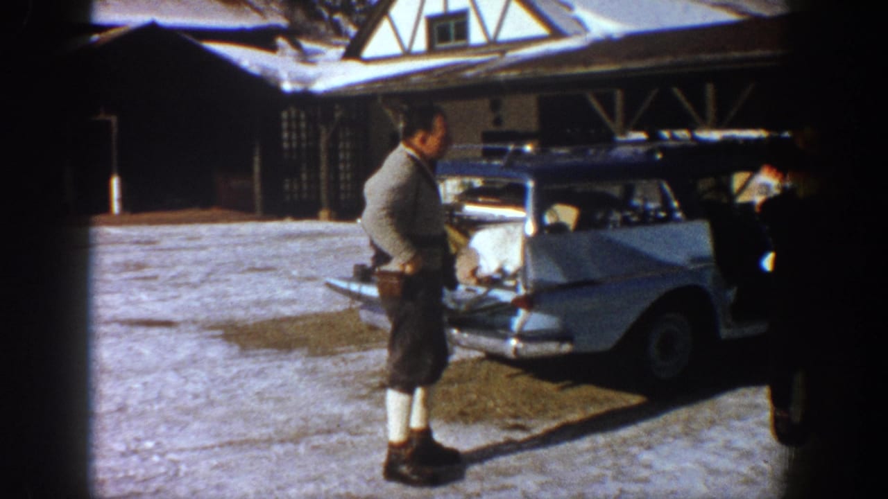 A lone man wearing shorts, knee-high socks, and a light sweater stands beside a pale 1960s American car; open timber-frame structure visible behind, Aspen area, 1961.