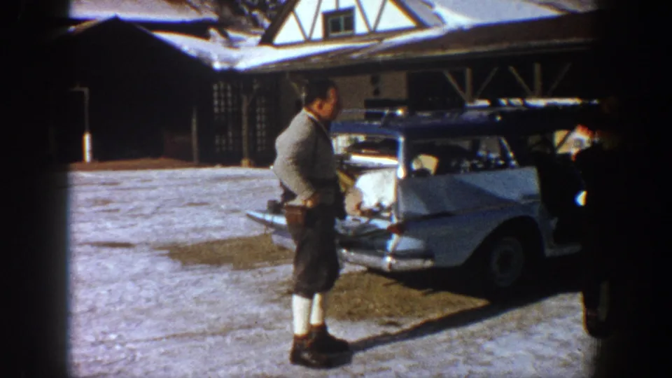 A lone man wearing shorts, knee-high socks, and a light sweater stands beside a pale 1960s American car; open timber-frame structure visible behind, Aspen area, 1961.