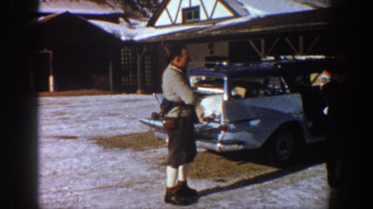 Man in knit sweater, shorts and white socks preparing ski gear at a pale 1960s station wagon, Tudor-style lodge behind, Aspen Colorado circa 1961.