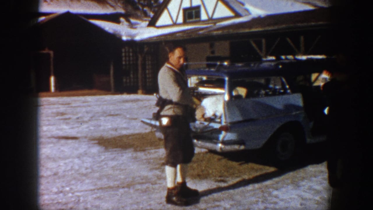 Man in ski clothing adjusting gear at a pale station wagon's open rear hatch, Tudor-style alpine lodge visible behind, Aspen Colorado circa 1961.
