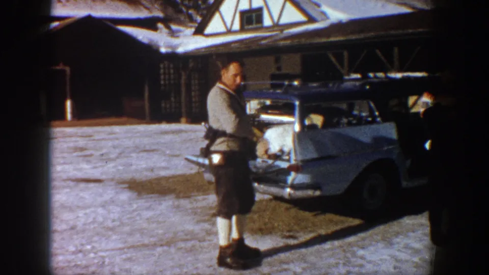 Man in ski clothing adjusting gear at a pale station wagon's open rear hatch, Tudor-style alpine lodge visible behind, Aspen Colorado circa 1961.