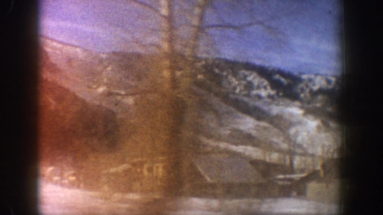 Opening frame of an 8mm home movie shot from a moving car, showing rustic buildings and a snow-covered mountain ridge consistent with Aspen, Colorado, 1961.