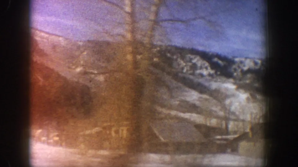 Opening frame of an 8mm home movie shot from a moving car, showing rustic buildings and a snow-covered mountain ridge consistent with Aspen, Colorado, 1961.