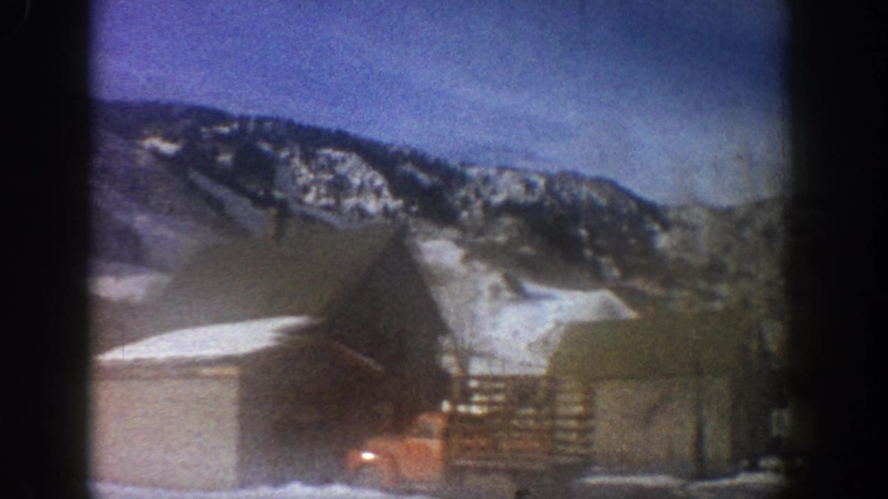 8mm home movie frame from a moving car showing a wooden barn with exterior stairs, a red vehicle, and a snow-covered mountain ridge, consistent with Aspen, Colorado, 1961.