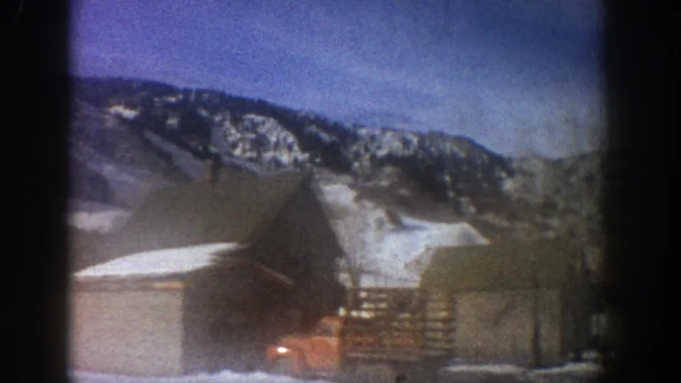 8mm home movie frame from a moving car showing a wooden barn with exterior stairs, a red vehicle, and a snow-covered mountain ridge, consistent with Aspen, Colorado, 1961.