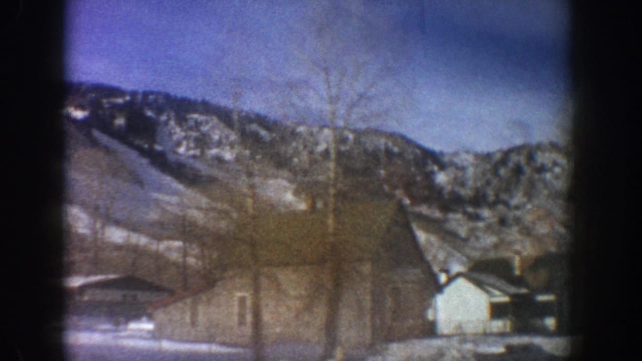 Closing 8mm frame with shifted car-window perspective showing a small white structure and bare trees against a snow-covered mountain ridge, consistent with Aspen, Colorado, 1961.