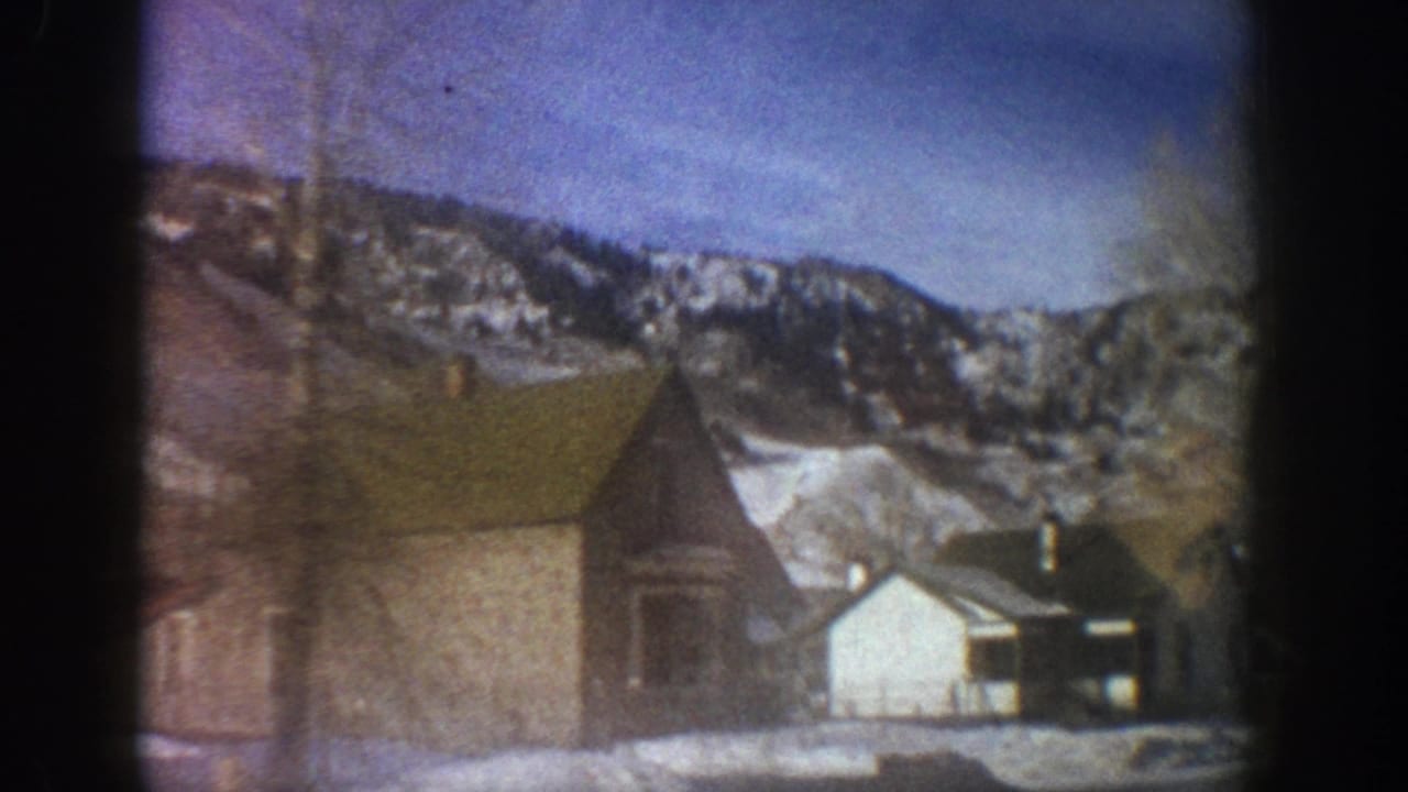 8mm home movie frame showing rustic wooden buildings in Aspen, Colorado winter valley, circa 1961, with snow-covered Rocky Mountain peaks in background.