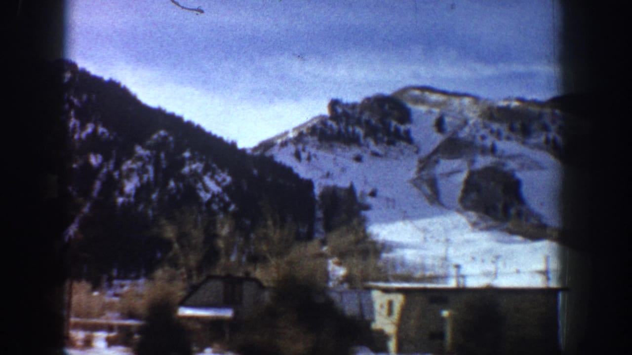 Snow-covered mountain range with rocky peaks above small valley structures, bare trees in midground, pale blue sky.