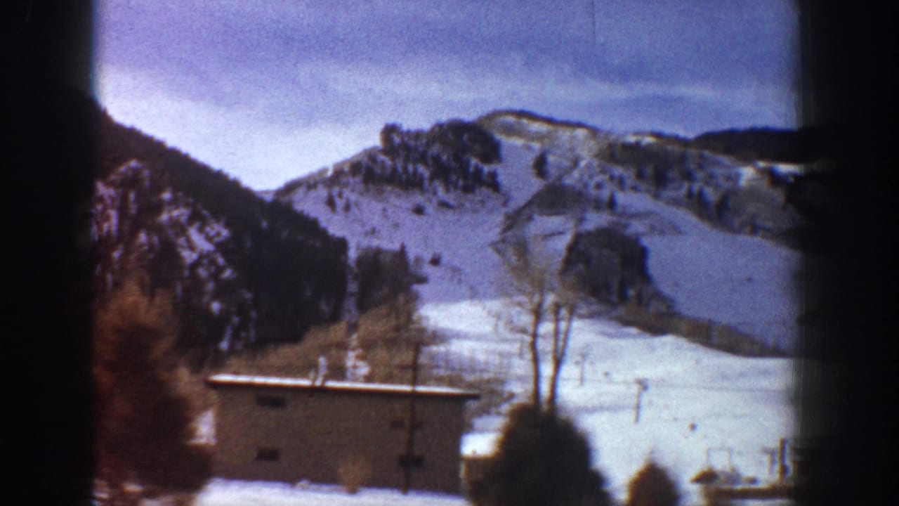 Color-faded 8mm frame showing broad snow-covered valley with small buildings and bare trees, mountain slopes above, consistent with 1961 Aspen, Colorado.