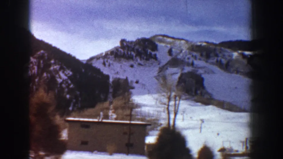 Color-faded 8mm frame showing broad snow-covered valley with small buildings and bare trees, mountain slopes above, consistent with 1961 Aspen, Colorado.