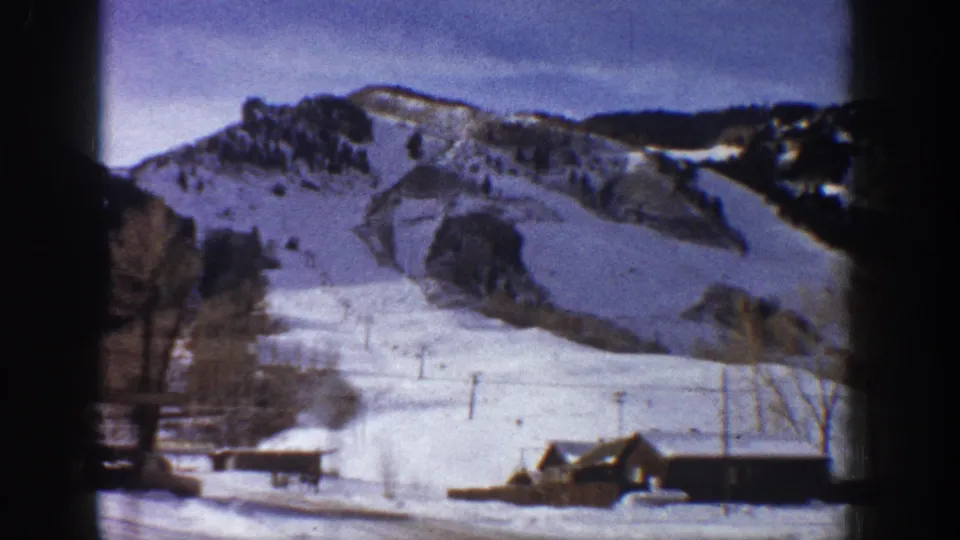 Continuing 8mm pan over snow-covered Aspen valley with small buildings and bare trees under winter sky, circa 1961.