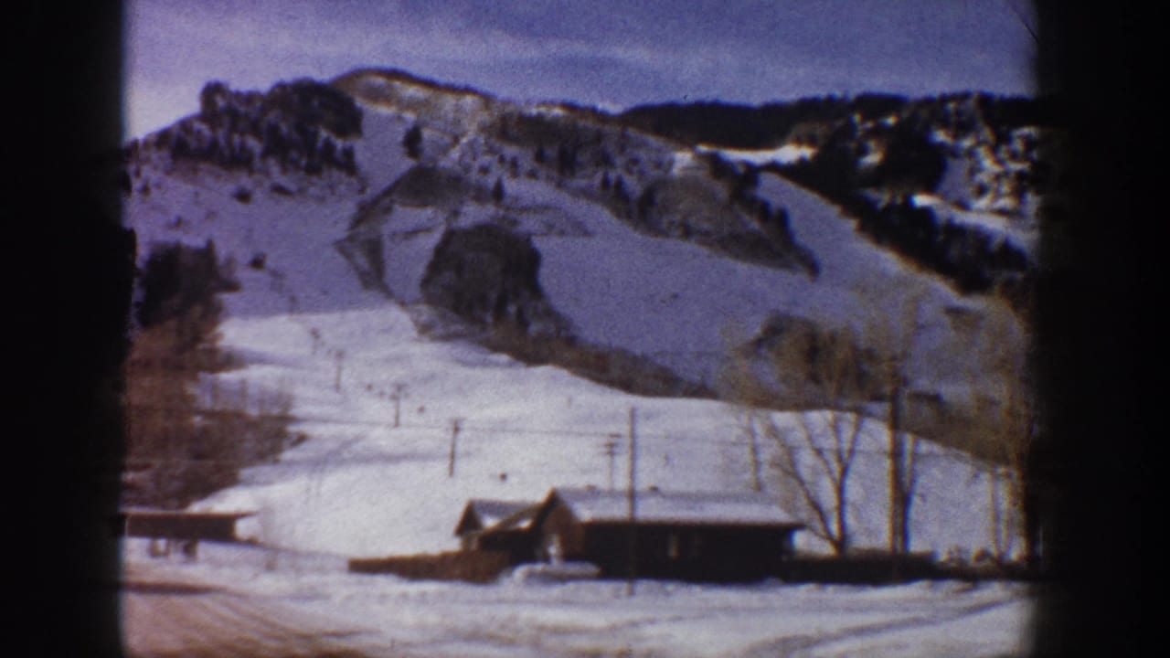 8mm home movie frame showing a cleared ski run, timber cabin, and bare trees against a snow-covered mountain in early 1960s Aspen, Colorado.