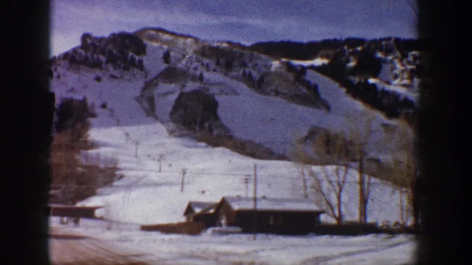 8mm home movie frame showing a cleared ski run, timber cabin, and bare trees against a snow-covered mountain in early 1960s Aspen, Colorado.