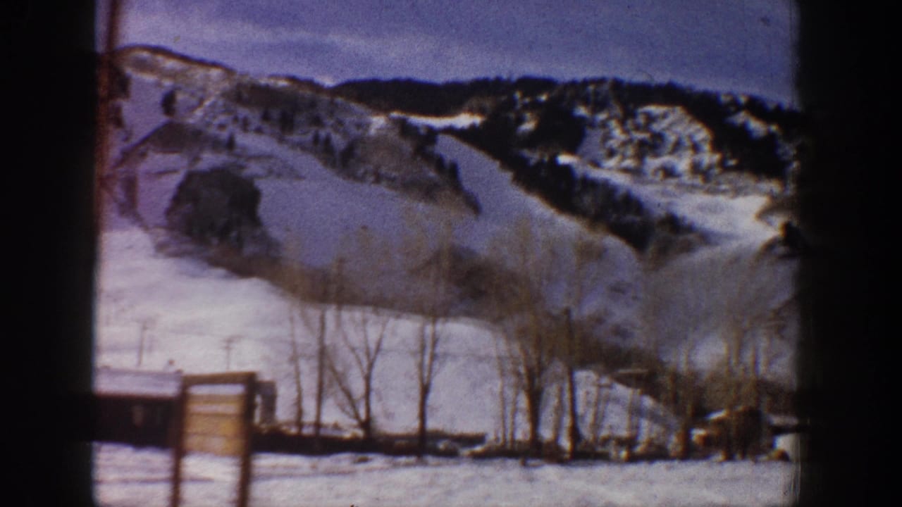 8mm home movie frame of a snowy ski mountain with a large wooden sign structure visible at the base and bare trees in the foreground.