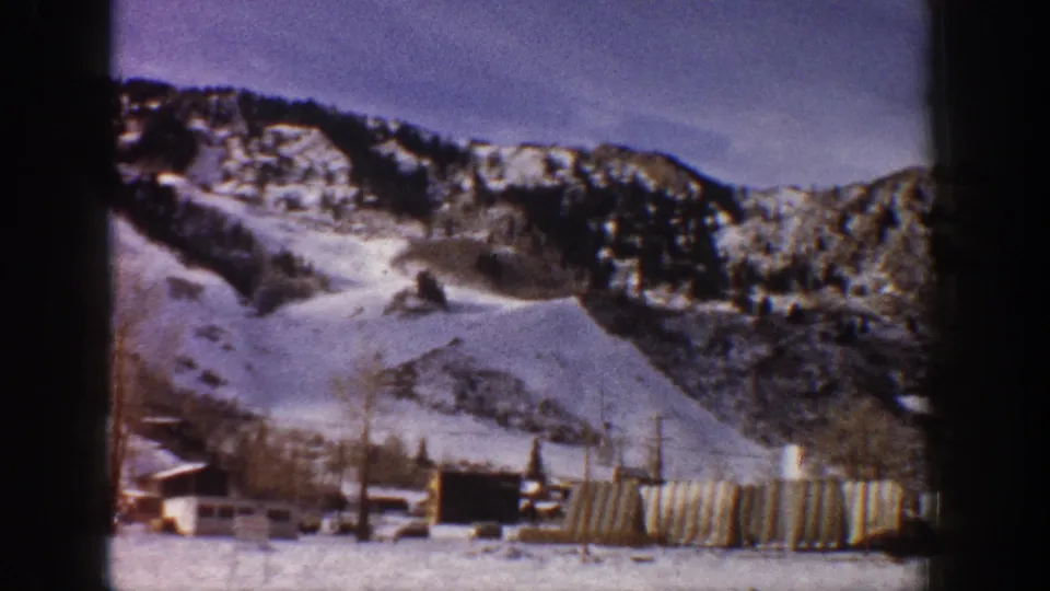 Wide 8mm home-movie shot of snow-covered mountain slopes and village buildings at base, consistent with early 1960s Aspen, Colorado.
