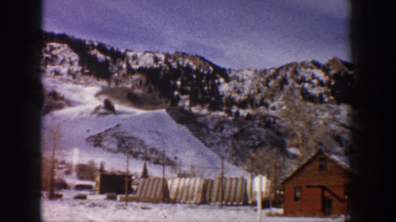 Similar wide shot of snow-covered mountain with village base; red-brown timber building visible at right edge; 8mm home movie.