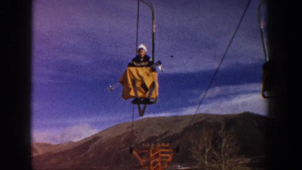 A rider in white winter hat ascends on a vintage single-seat chairlift with yellow cover at what appears to be a Colorado ski resort, circa 1961, filmed on 8mm.